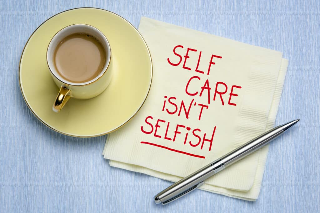 Cream napkin on blue table with words "self care is not selfish" written on it in red writing. A pen sits by the side and next to a yellow cup and saucer, filled with tea - Julia Ngapo Business Coaching